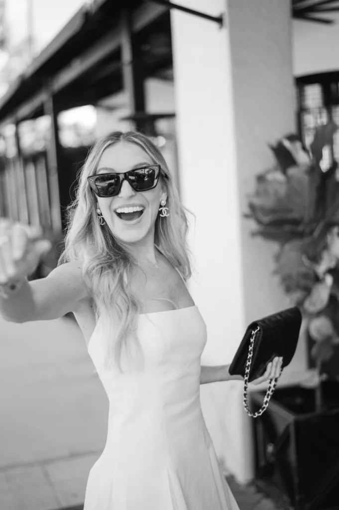 Joyful bride in a chic white dress and stylish sunglasses reaching out in a playful greeting, captured by renowned Tampa wedding photographer Hernan in Naples, Florida