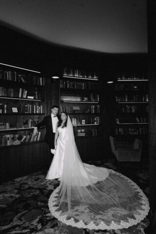 Bride and groom pose elegantly in the Hotel Haya library, showcasing the bride’s long, lace-trimmed veil